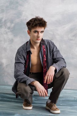 Düğmeleri iliklenmemiş gömlekli ve kravatlı genç adam gri arka planda çömelmiş, ciddi bir ifadeyle kameraya bakıyor..