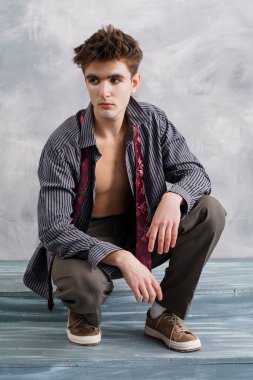 Kısmen düğmesiz çizgili gömleği ve kravatıyla çömelmiş genç adam, dokulu bir arka planda doğrudan izleyiciye bakıyor..