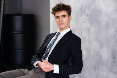 Koyu renk takım elbiseli ve kravatlı genç adam kapalı bir yerde oturmuş, gri bir arka planda kameraya bakıyor..