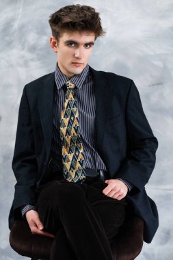 Kapalı bir yerde oturan, koyu renk bir takım giyen, desenli kravat takan, çizgili gömlek giyen ve doğrudan izleyiciye bakan resmi kıyafetli genç bir adam..