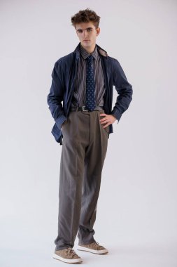 Şık giyinmiş genç bir adam, beyaz bir arka plan karşısında duruyor. Ceket, gömlek, kravat ve pantolon giyiyor..