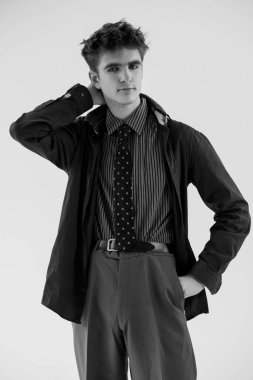 Resmi giyinmiş şık bir genç adam, beyaz bir arka planda el ele poz veriyor, klasik moda ve kendine güvenen tavırlar sergiliyor..