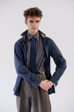 Sade beyaz bir arka plana poz veren şık giyinmiş genç bir adam. Donanma ceketi, çizgili gömlek ve kahverengi pantolonlu mavi bir kravat takıyor..