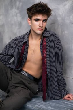 Kısmen düğmesiz gömlekli ve gevşek kravatlı genç bir adam, gri bir zemine karşı poz veriyor, kameraya ciddi bir ifadeyle bakıyor..