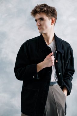 Tarz sahibi genç bir adam beyaz tişört ve gri pantolonun üzerine siyah ceket giyip açık renk arka planda poz veriyor..