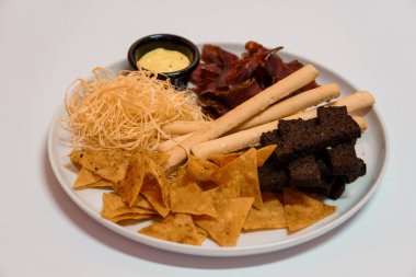 Tortilla cipsi, ekmek çubuğu, kurutulmuş et, melek saçı makarna ve beyaz arka planda siyah kasede sarı soslu çeşitli atıştırmalıklar..