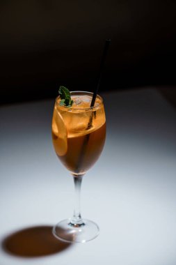 Portakal dilimi ve nane kokteyle sarılı taze bir kokteyl, stüdyo ışığı altında beyaz yüzey üzerinde siyah pipetle servis edilir..