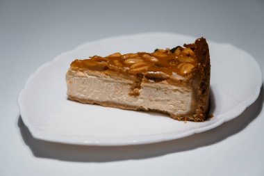 Beyaz tabakta nefis bir dilim fıstık karameli cheesecake, yumuşak ışıklandırmalı stüdyo fotoğrafı, zengin dokular ve davetkar tatlı sunumu..