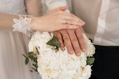 Bir gelin ve damadın elini gösteren yakın plan bir düğün fotoğrafı. Bir buket beyaz gülün üzerinde dinlenen alyanslar aşkı, bağlılığı ve yeni bir başlangıcı simgeliyor..