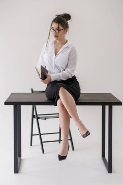 Masada oturmuş kitap okuyan genç bir iş kadını, profesyonel kıyafetler, odaklanmış ifade, ofis dekorasyonu, siyah etek..