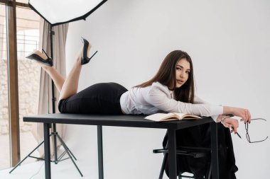 Kendine güvenen genç iş kadını, açık bir kitap ve gözlük ile ofis masasında yatıyor, şık kıyafetler, stüdyo fotoğrafı..