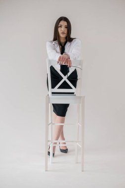 Tarafsız bir arkaplana karşı beyaz sandalyede poz veren iş elbiseli zarif bir kadın..