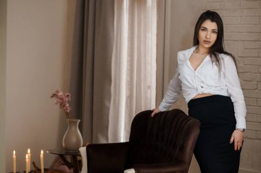 Beyaz gömlekli ve siyah etekli zarif bir kadın. Sandalyeye yaslanmış, içinde mumlar olan tuğladan bir iç mekan..