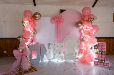 Bir balon kemeri, bir tek işareti ve bir sandalye ve pasta eşliğinde prenses temalı bir arka plan içeren büyüleyici bir pembe ve altın rengi ilk doğum günü partisi..
