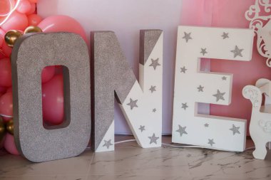 Göz kamaştırıcı bir ilk doğum günü kutlaması arka planında büyük, pırıl pırıl, yıldızlarla süslenmiş tek harfli süslemeler, yanında pastel pembe bir balon çelengi ve tek boynuzlu at heykeli..