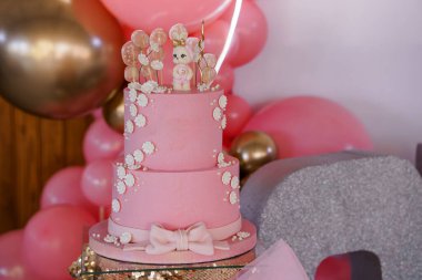 Sevimli bir tavşan figürü ve beyaz çiçek aksanıyla süslenmiş iki katlı sevimli pembe bir doğum günü pastası. Balon kemeri ve altın süslemelerle süslenmiş..
