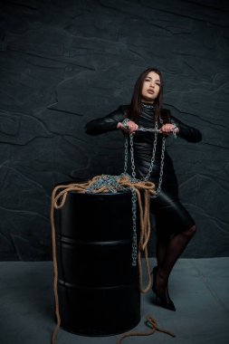 Siyah deri ceketli genç bir Asyalı kadın, koyu renk, dokulu bir stüdyo ortamında bir yağ varilinin yanında zincirlerle ve halatlarla dramatik bir şekilde poz veriyor..