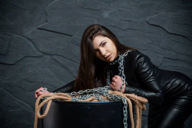 Siyah deri elbiseli çarpıcı bir kadın, zincirlenmiş ve çarpıcı bir şekilde koyu gri bir duvara karşı poz vermiş, esaret ve kırılganlık temalarını somutlaştırmış..