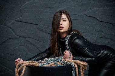 Uzun kahverengi saçlı büyüleyici genç bir kadın koyu renkli, desenli bir ortamda, deri bir kıyafet giymiş ve zincir ve iple süslenmiş bir şekilde dramatik bir şekilde poz veriyor..