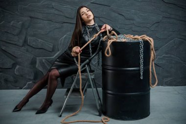 Siyah deri elbiseli çarpıcı genç bir kadın, güçlü ve güçlü temaları somutlaştıran, koyu taş bir duvara karşı halat ve zincirlerle çarpıcı bir poda poz veriyor..