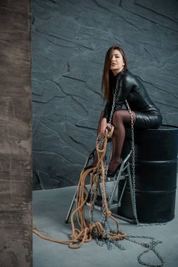 Uzun kahverengi saçlı, siyah deri bir elbise ve yüksek topuklu ayakkabı giyen beyaz bir kadın, metal bir fıçının üzerinde oturuyor..