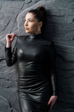 Göz alıcı, zeytin tenli, parlak siyah deri bir balıkçı yaka elbise ve altın küpeler giyen genç bir kadın, koyu gri gri bir duvara karşı güven içinde poz veriyor..