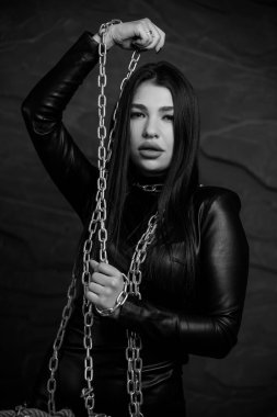Siyah deri kıyafetli genç bir Doğu Asyalı kadının dramatik portresi. Elinde gümüş bir zincir var. Arkaplanda koyu renk bir zemin var..