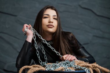 Uzun siyah saçlı ve dolgun dudaklı büyüleyici genç bir kadın gümüş bir zincir tutuyor, koyu gri bir duvara karşı kalın bir iple poz veriyor, kendinden emin..