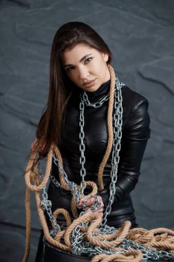 Uzun siyah saçlı, siyah deri kıyafetli, kalın iplerle ve gümüş zincirlerle bezenmiş bir kadının dramatik portresi..