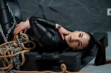 Açık tenli çarpıcı bir kadın, zayıf bir stüdyo ortamında halat ve zincirlerle bağlanmış ahşap sandıkların üzerinde tahrik edici bir şekilde yatıyor..