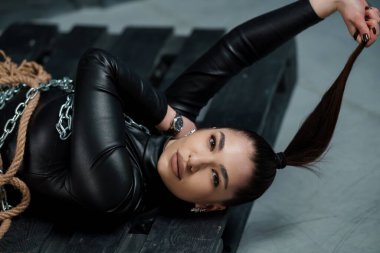 Zarif bir at kuyruğu ve deri bluzu olan çarpıcı bir kadın ahşap bir palette zincirlerle güven içinde yatıyor, loş bir endüstriyel ortamda güçlü ve asi bir estetik yayıyor..