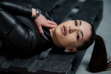 Çekici, kahverengi saçlı beyaz bir kadın koyu ahşap bir bankta tahrik edici bir şekilde uzanıyor. İnce, çekici bir ifadeyle kameraya bakıyor. Siyah deri ceket giyiyor ve lüks bir saat sergiliyor..