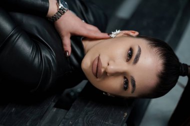 Ahşap bir yüzeye uzanmış kusursuz makyajlı bir kadının çarpıcı portresi, lüks bir stil sergiliyor deri kıyafetler ve zarif mücevherler, koyu estetik.