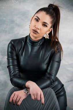 Siyah deri kıyafetli büyüleyici bir kadın. Gri desenli arka planı olan bir stüdyo ortamında özgüvenle poz veriyor. Parlak bir saç stili ve altın mücevherler sergiliyor..