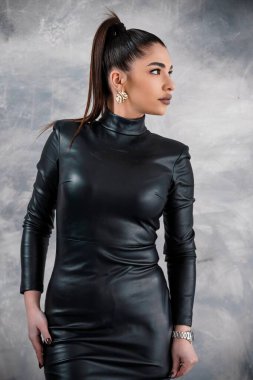 Parlak at kuyruğu ve kalın altın küpeli çarpıcı bir kadın, tarafsız gri bir arkaplana karşı modern ve sofistike bir stil sergileyen siyah deri bir elbise giyiyor..