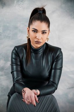 Siyah saçlı, uzun at kuyruklu, parlak siyah deri elbiseli ve altın küpeli bir kadının dikkat çekici portresi..