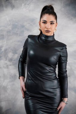 Gösterişli siyah deri elbiseli şık bir kadın tarafsız gri bir arkaplana karşı güvenle poz veriyor, modern, sofistike bir estetik, cesur mücevherler ve dramatik bir saç stili sergiliyor..