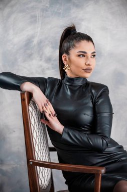 Tahta bir sandalyeye oturmuş kendine güvenen bir bakışla çarpıcı bir kadın, siyah deri bir elbise ve altın küpeler takıyor tarafsız gri bir arka plana karşı..