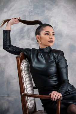 Parlak at kuyruklu genç bir kadının büyüleyici bir portresi. Ahşap bir sandalyeye oturmuş siyah deri bir elbiseyle poz veriyor..