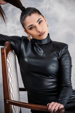 Uzun siyah saçlı, uzun, at kuyruklu çarpıcı bir genç kadın. Gri dokulu bir duvara karşı, parlak, siyah bir deri elbiseyle poz veriyor..