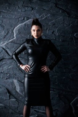Kendine güvenen, parlak siyah deri elbiseli bir kadının dramatik portresi. Koyu dokulu taş bir duvara karşı poz veriyor. Güçlü ve sofistike..