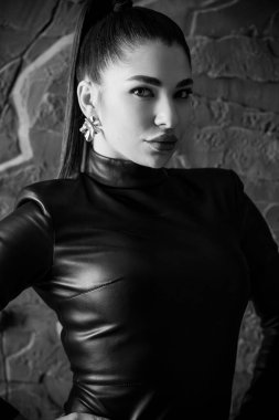 Çarpıcı bir siyah beyaz portre, Hailey Bieber, yüksek at kuyruklu genç bir kadın, siyah deri bir elbise ve gümüş küpeler giyiyor, güvenle poz veriyor..