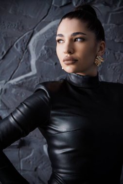 Siyah deri elbisesi ve altın küpeleri olan, uzun topuz şeklinde siyah saçlı, düşünceli bir genç kadının çarpıcı portresi..
