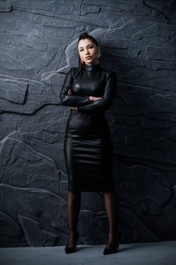 Gösterişli, siyah deri elbiseli, kolları çapraz, dokulu, taş bir zemin üzerinde duran genç bir İspanyol kadının dramatik portresi..