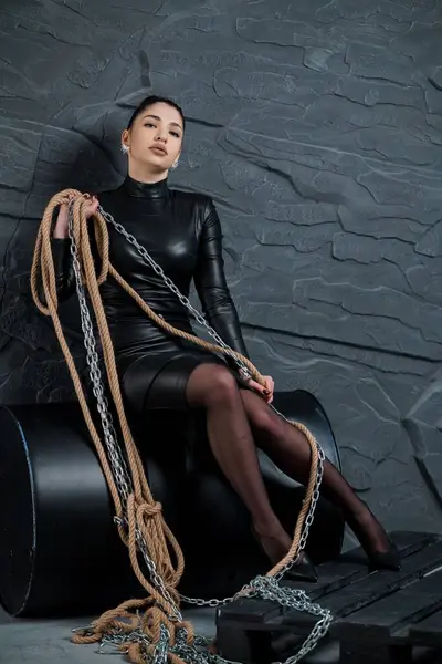 Siyah lateks bir elbise içindeki siyah ve büyüleyici bir kadın portresi modern bir sandalyeye oturmuş halatlar ve zincirlerle, somut bir güç ve gizem ile..