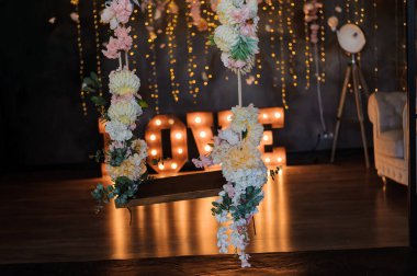 Romantik düğün arka planında, beyaz ve pembe çiçeklerden oluşan kademeli çiçek aranjmanlarıyla süslenmiş büyük, aydınlık bir aşk işareti ve loş bir iç mekan var..