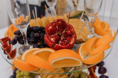 Portakallar, narlar, üzümler, incirler ve şarap kadehleriyle beyaz masa ortamında zarif bir meyve tabağı düzenleme, şenlikli ve lüks bir atmosfer yaratıyor..