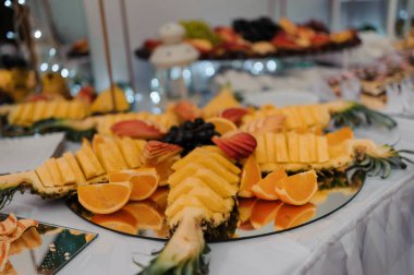 Dilimlenmiş mangolar, portakallar, ananas, üzümler ve sanatsal olarak dizilmiş diğer renkli meyvelerin yer aldığı bir etkinlikte aynalı bir yüzey üzerinde bol ve canlı bir meyve tabağı sunumu.