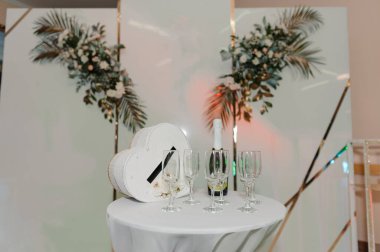 Beyaz Çiçek Düzenlemeleri ve Çembersel Masa Üzerinde Şampanya Flütleri ile Şık Düğün Resepsiyonu Altın Aksanlı Modern Beyaz Arkaplana Karşı.