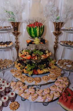 Altın aksanlı ve beyaz kumaşlarla süslenmiş meyve düzenlemeleri, pastalar ve pastaların yer aldığı bir düğün resepsiyonunda ayrıntılı bir tatlı büfesi..